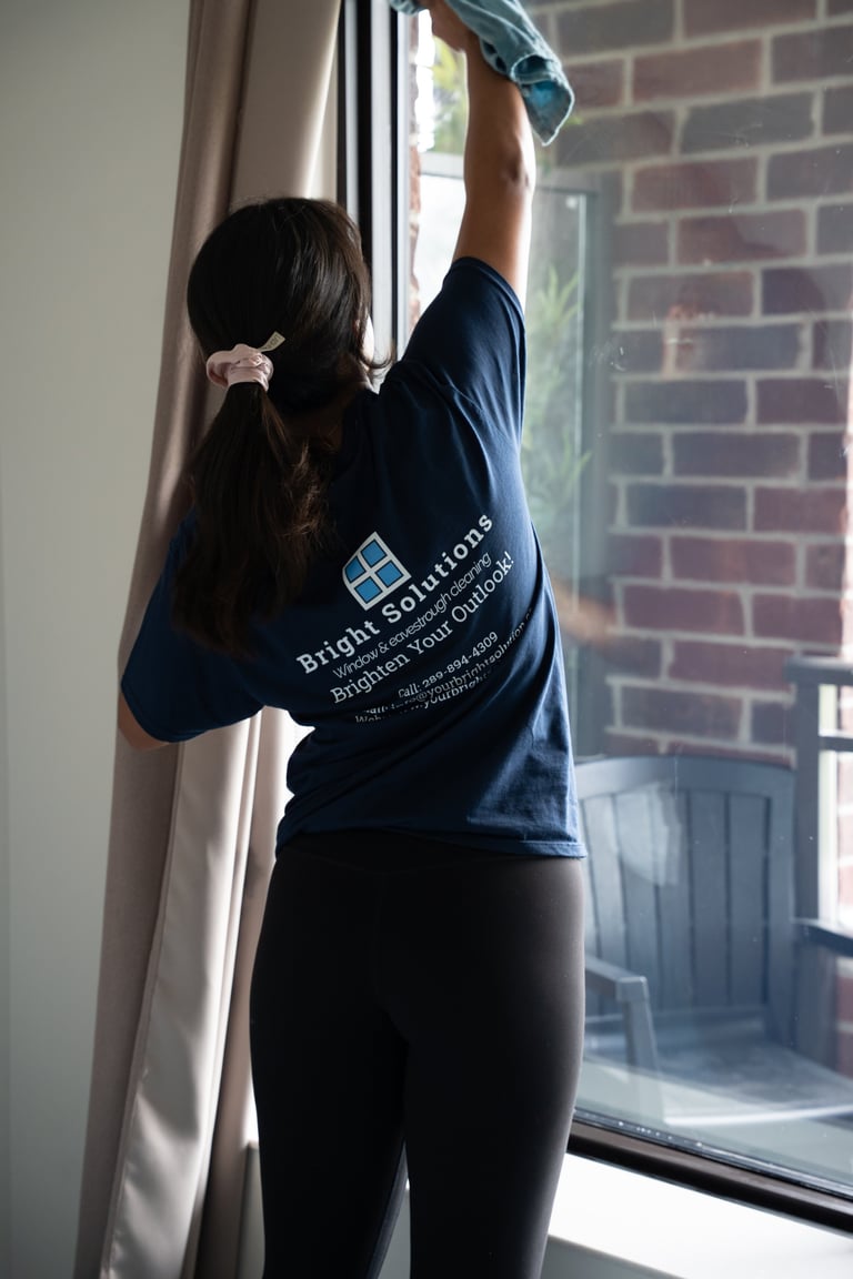 Person in navy shirt cleaning a window with a cloth, standing indoors with brick wall visible outside
