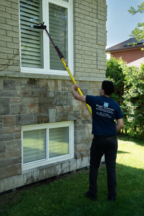 Worker in navy shirt using long pole to clean exterior wall of stone house with white windows