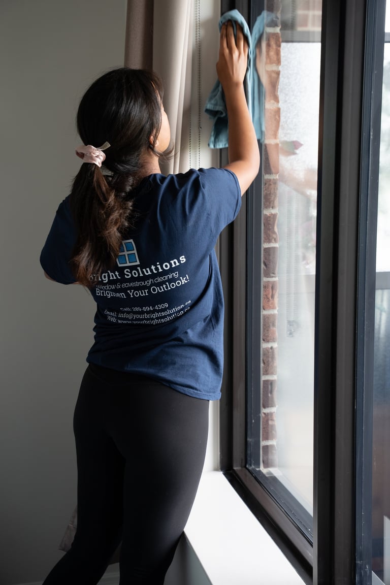 Person in navy Brick Solutions shirt cleaning a window with a microfiber cloth, brick exterior visible outside